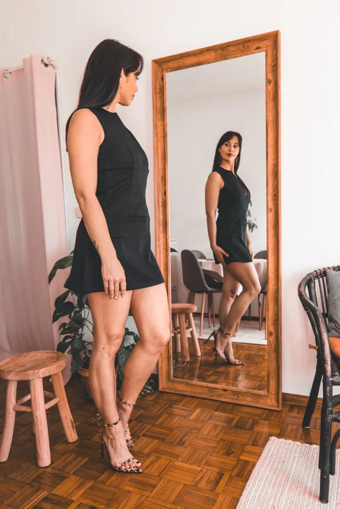 Caroline en robe noire élégante et talons PrettyLittleThings, se regarde dans un miroir, ambiance chic et moderne.