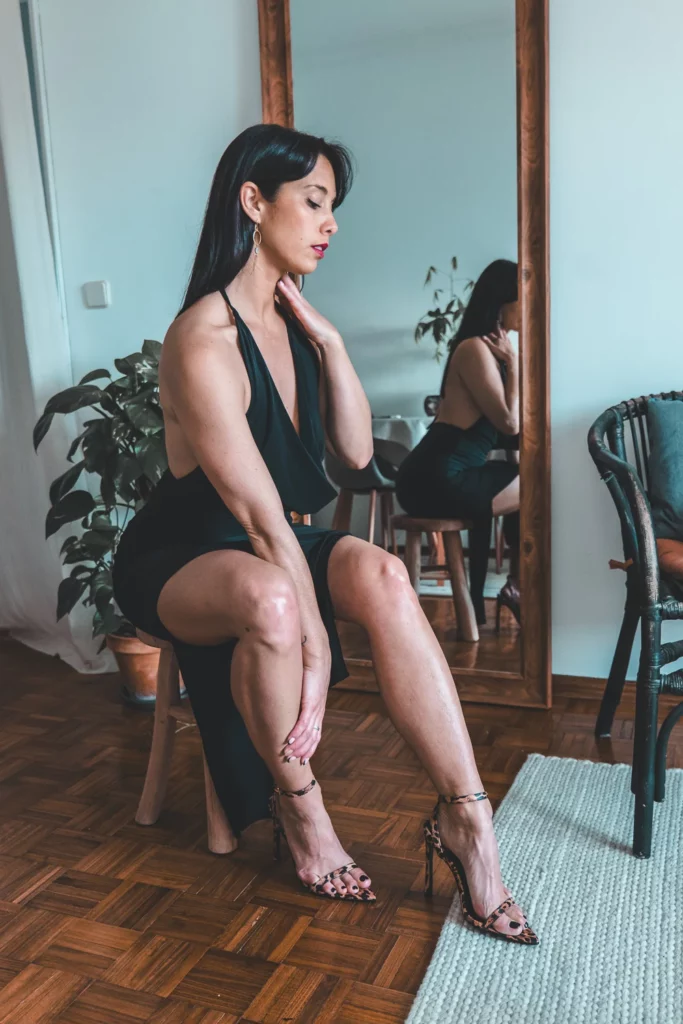 Caroline en robe noire élégante PrettyLittleThings et talons, posant devant un miroir dans un intérieur moderne.