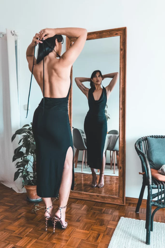 Caroline ajuste sa robe noire dos nu PrettyLittleThings devant un miroir, talons assortis, ambiance élégante et moderne.