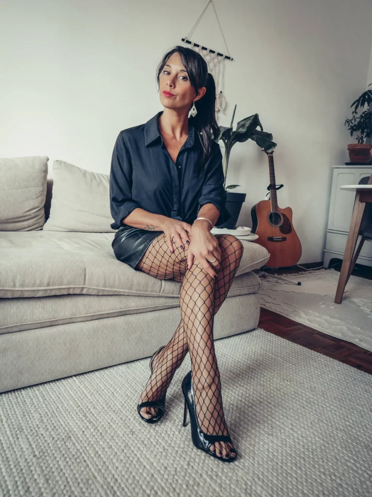 Caroline assise sur un canapé beige, vêtue d'une chemise bleu marine, d'une jupe en cuir noir et de collants filet noirs.