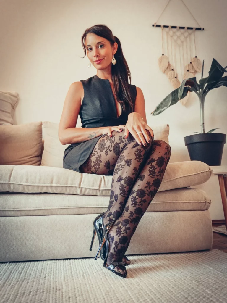 Caroline assise sur un canapé beige, portant une robe noire sans manches et des collants à motifs floraux dans un salon en Algarve.