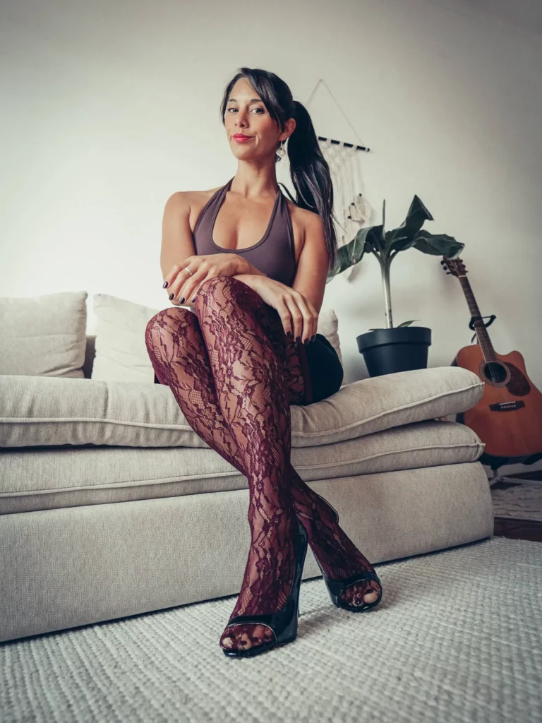 Caroline assise sur un canapé beige, vêtue d'un haut en halter marron, de collants en dentelle bordeaux et de chaussures à talons noirs, dans un intérieur avec plante et guitare.