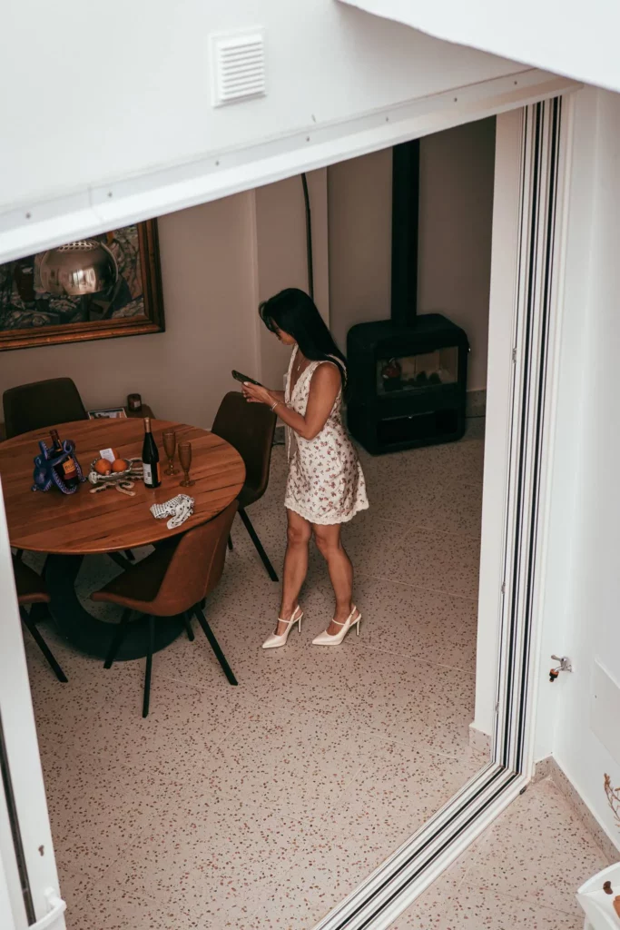 Caroline debout dans une salle à manger, vêtue d'une robe fleurie et de talons hauts, tenant un téléphone. La pièce est équipée d'une table ronde, d'une cheminée et de détails décoratifs.