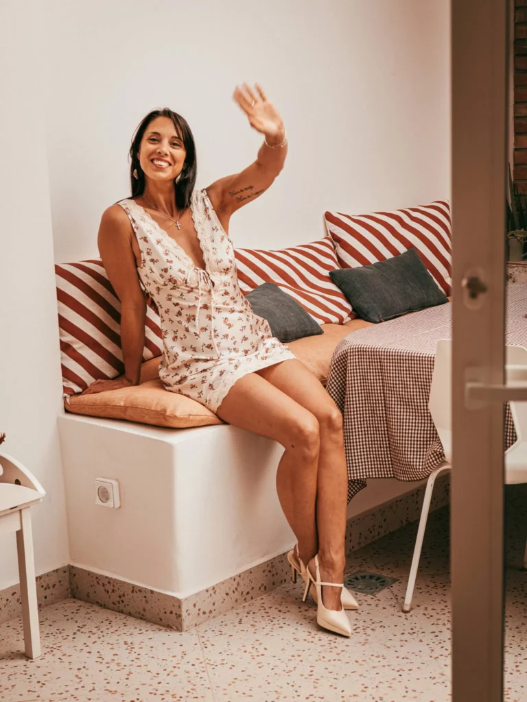 Caroline assise sur un banc avec des coussins rayés, vêtue d'une robe fleurie et de chaussures à talons.