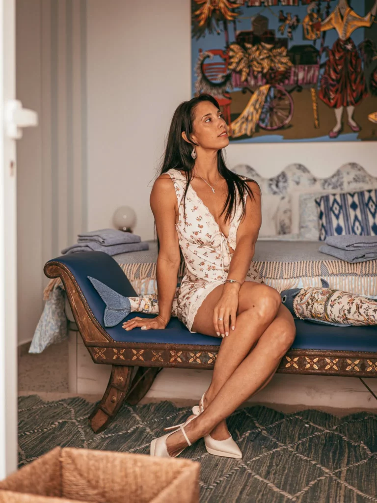 Caroline assise sur un banc bleu dans une pièce décorée d'un lit avec coussins bleu-blanc et d'une grande peinture murale colorée. Elle porte une robe courte à imprimé floral et des escarpins blancs.