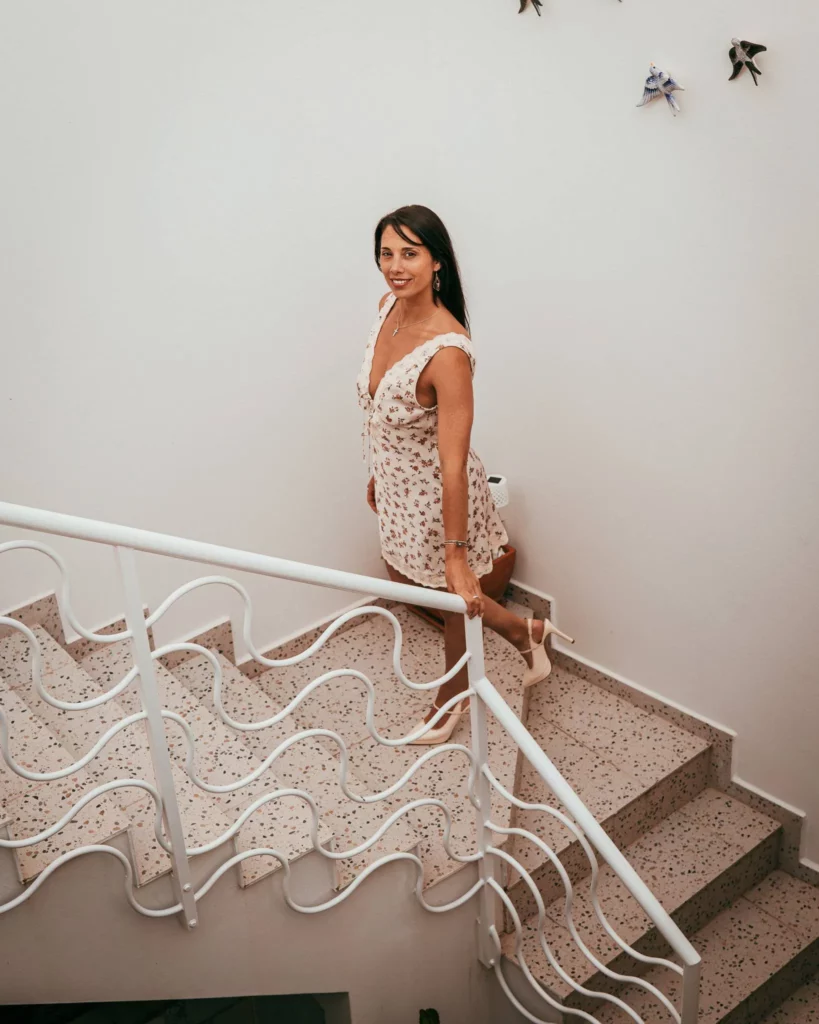 Caroline descend un escalier intérieur avec une robe à motifs floraux et des escarpins, dans un cadre éclairé par une lumière naturelle douce.