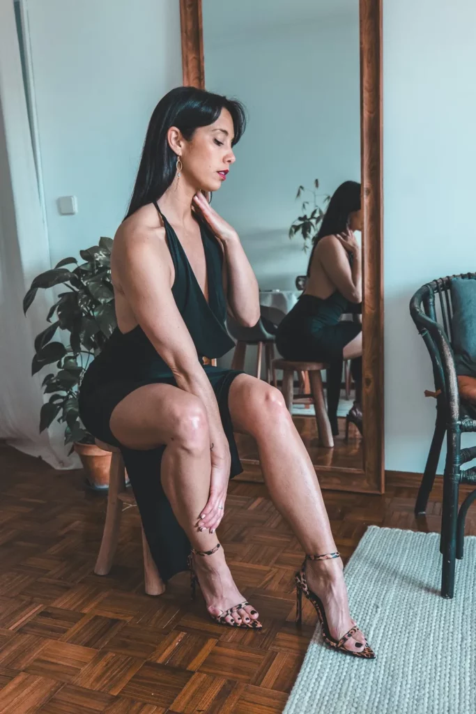Caroline est assise sur un tabouret, genou plié et main posée sur le cou, avec jambes étendues et une robe noire à décolleté profond.