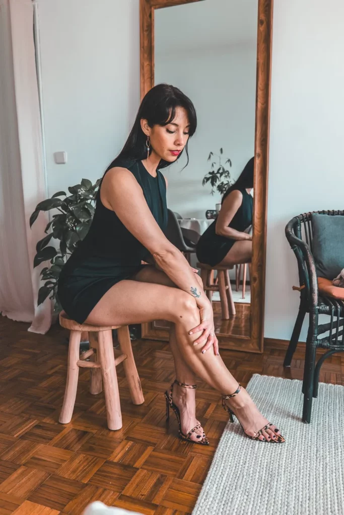 Caroline assise sur un tabouret en bois, portant une robe courte noire Pretty Little Thing et des talons à motifs, dans un intérieur lumineux avec un grand miroir et des plantes.