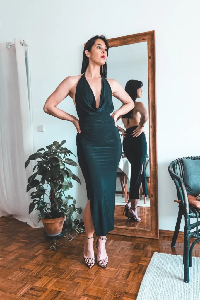 Caroline debout avec les mains sur les hanches, vêtue d'une robe noire ajustée. Elle se tient devant un grand miroir dans une pièce aux sols en parquet.