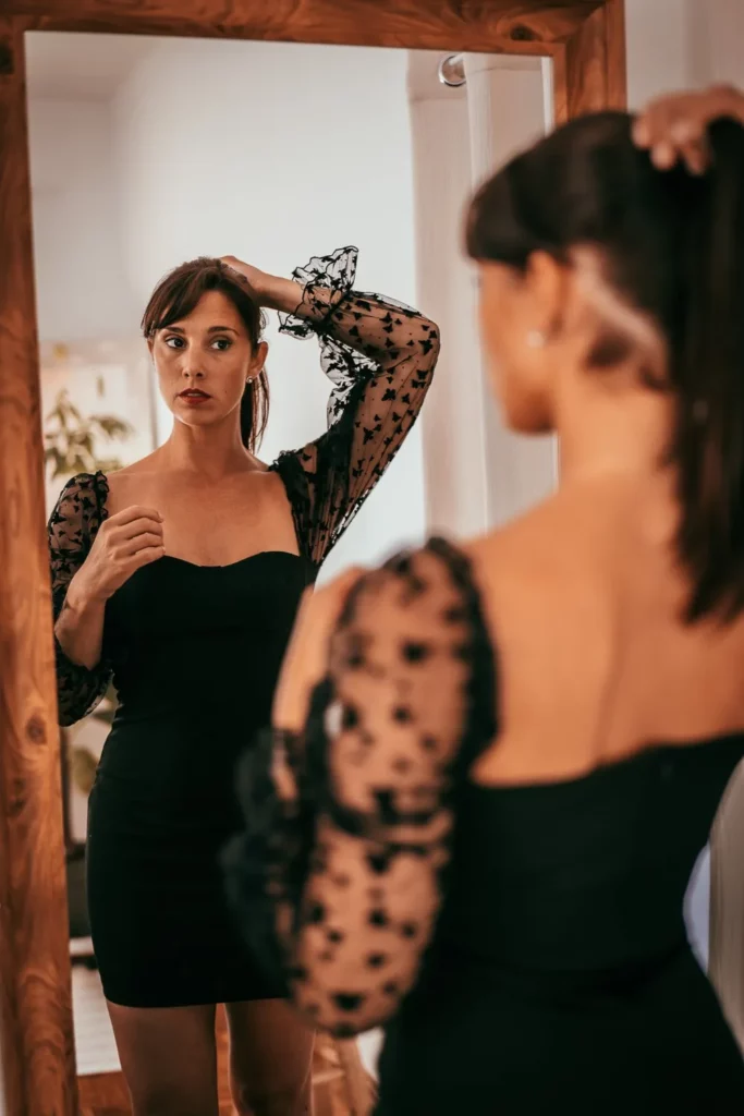 Caroline ajuste sa coiffure devant un miroir, portant une robe noire à manches longues en tulle à motifs dans un intérieur lumineux.