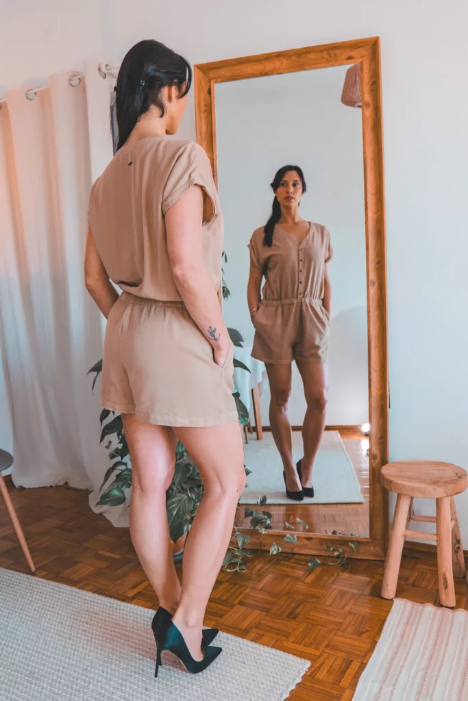 Caroline en combinaison beige et talons noirs se tient devant un grand miroir dans un intérieur lumineux, ambiance épurée typique d'un shooting Sunfere en janvier 2025.
