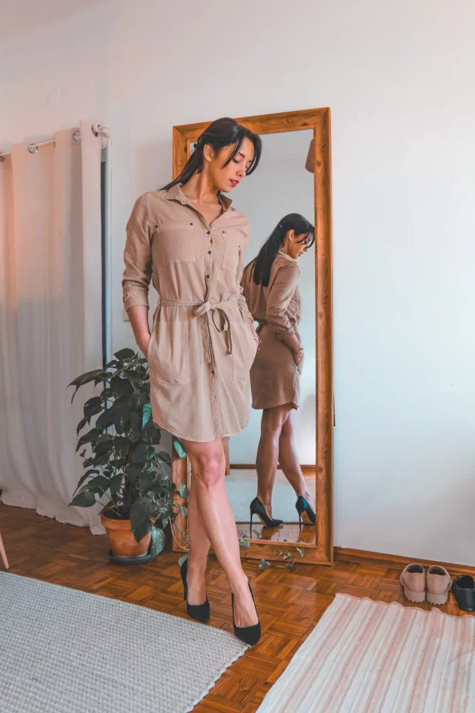 Caroline porte une robe beige et des escarpins noirs dans un intérieur lumineux, se tenant devant un miroir en bois. Une plante verte et des chaussures sont visibles au sol.