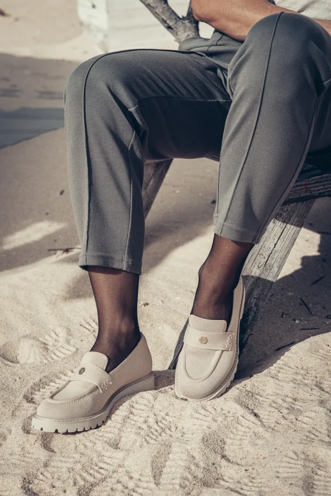 Gros plan sur les jambes d'une personne assise sur un banc en bois sur une plage de sable, portant un pantalon gris et des mocassins beiges. Ambiance ensoleillée typique de l'Algarve en février.