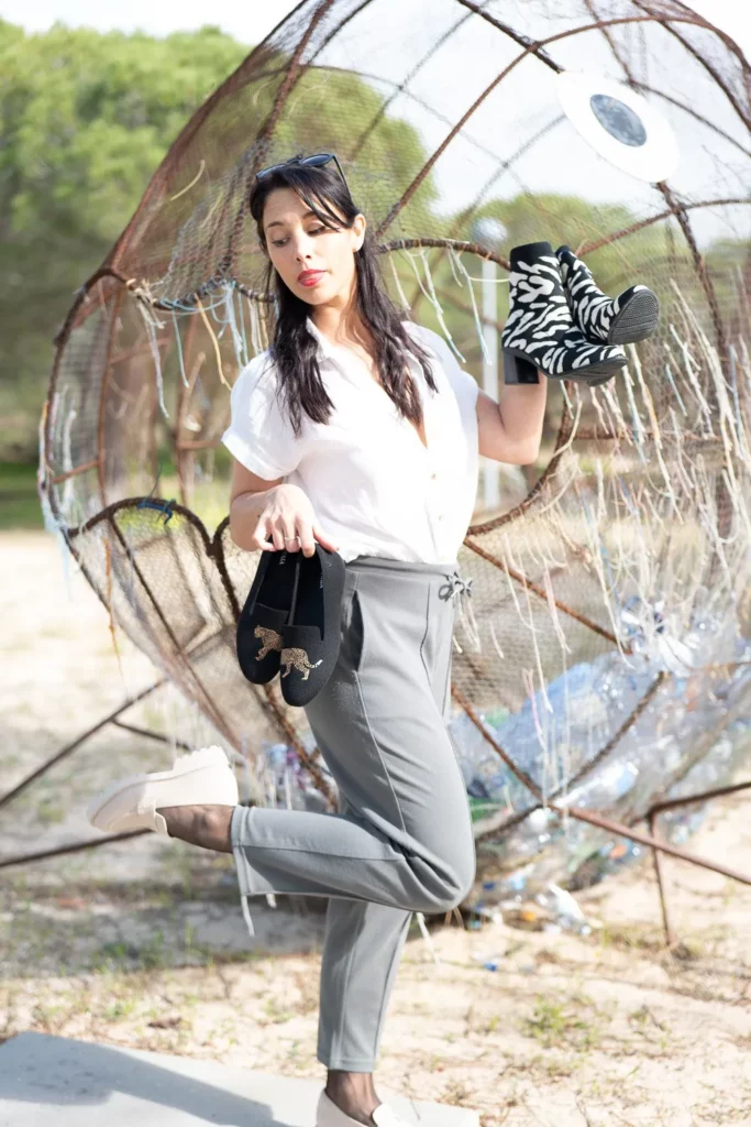 Caroline pose devant une sculpture métallique en plein air à Tavira, Algarve, en tenant deux paires de chaussures différentes. La scène est lumineuse et décontractée, avec une ambiance artistique en extérieur.