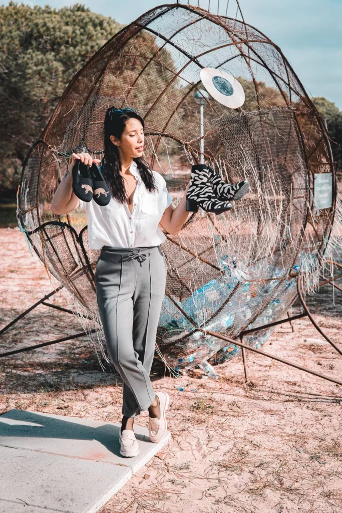 Caroline pose en extérieur devant une grande structure métallique artistique, tenant une paire de sandales noires et une paire de bottines zébrées. La scène se déroule sous un ciel clair, sur un sol sablonneux avec des arbres en arrière-plan.