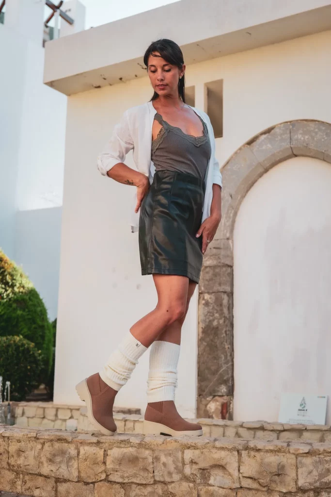Caroline pose devant une bâtisse blanche typique de l'Algarve, vêtue d'une jupe en cuir, d'un débardeur gris et de bottines beiges avec des jambières blanches. La lumière douce évoque une ambiance de fin d'après-midi en Algarve en février.