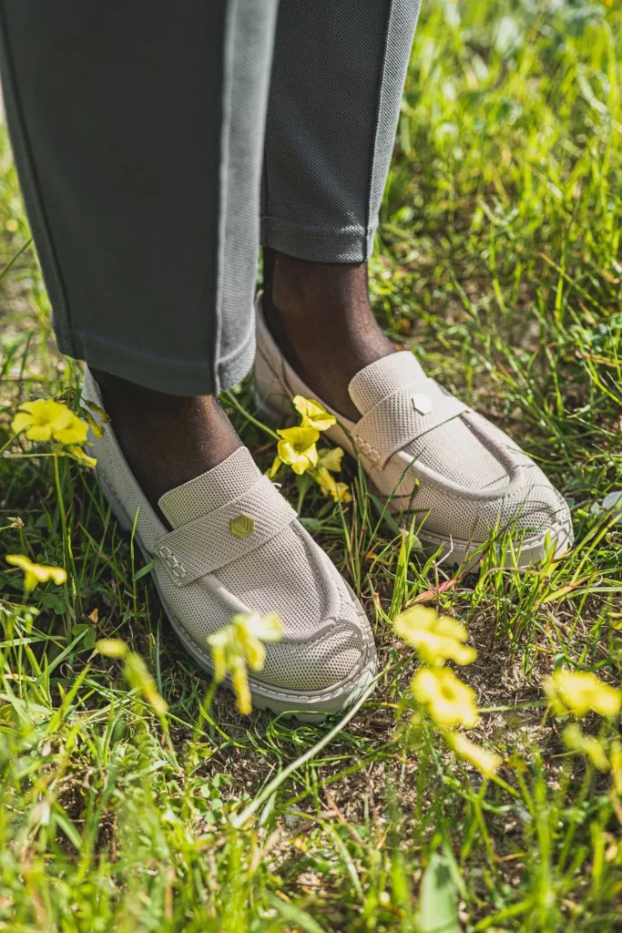Gros plan sur des chaussures beiges portées dans l'herbe parmi des fleurs jaunes, lumière naturelle en extérieur.