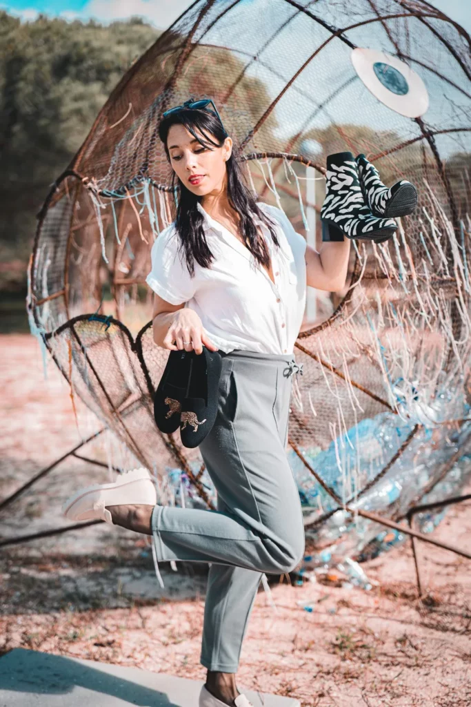 Caroline pose en extérieur à Tavira, Algarve, tenant deux paires de chaussures devant une grande structure artistique. La lumière et l'ambiance suggèrent un shooting mode en février.