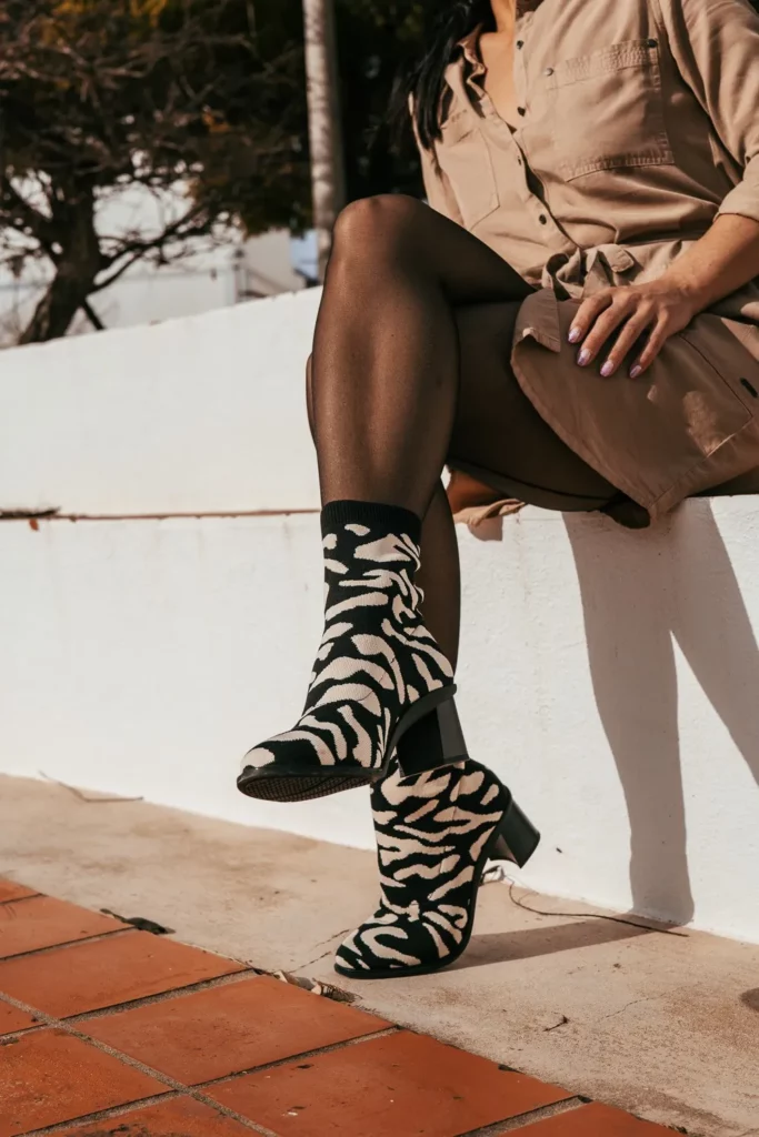 Caroline assise sur un muret blanc en extérieur à Tavira, Algarve, portant une robe beige et des bottines à motif zèbre. Lumière naturelle de février.