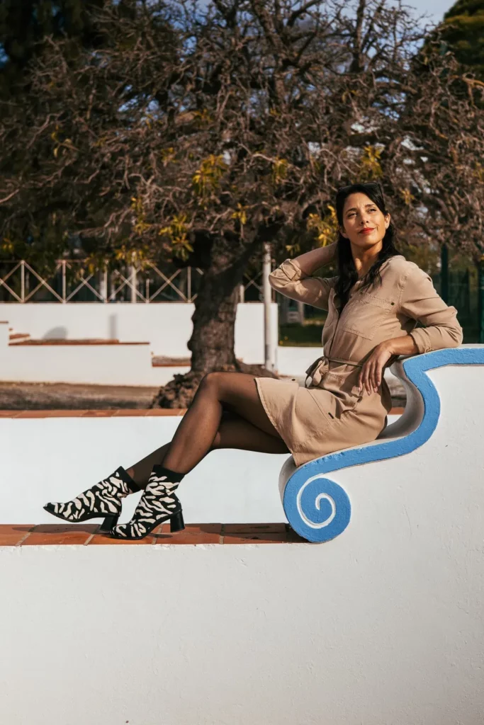 Caroline assise sur un banc blanc aux motifs bleus, en extérieur à Tavira dans l'Algarve, en février, portant une robe beige et des bottines à motif zèbre.